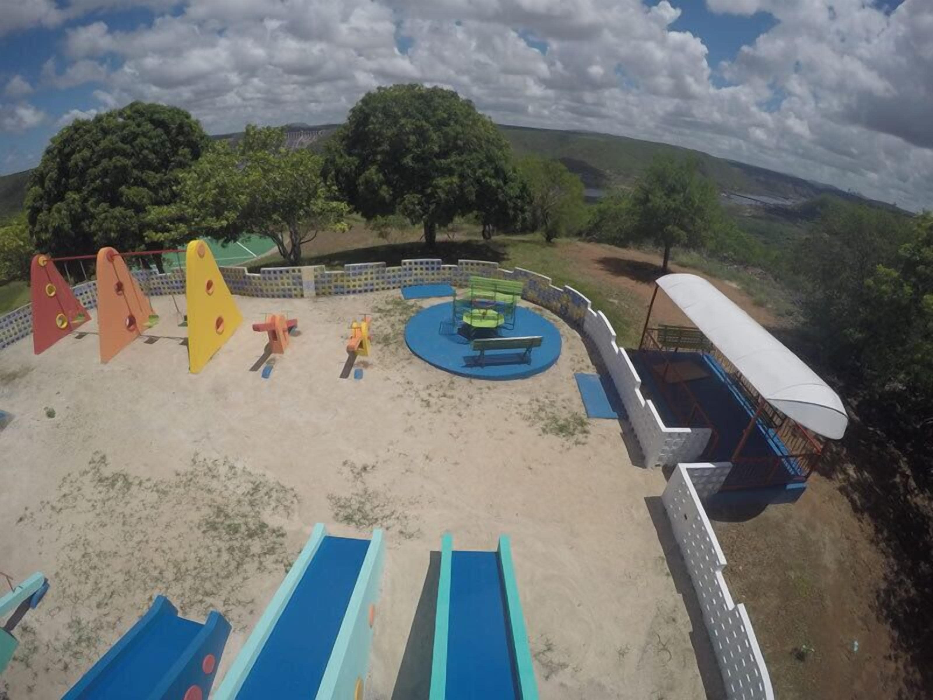 Parque infantil com escorregadores e baloiços, rodeado por um muro branco e árvores.