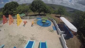 Parque infantil com escorregadores e baloiços, rodeado por um muro branco e árvores.