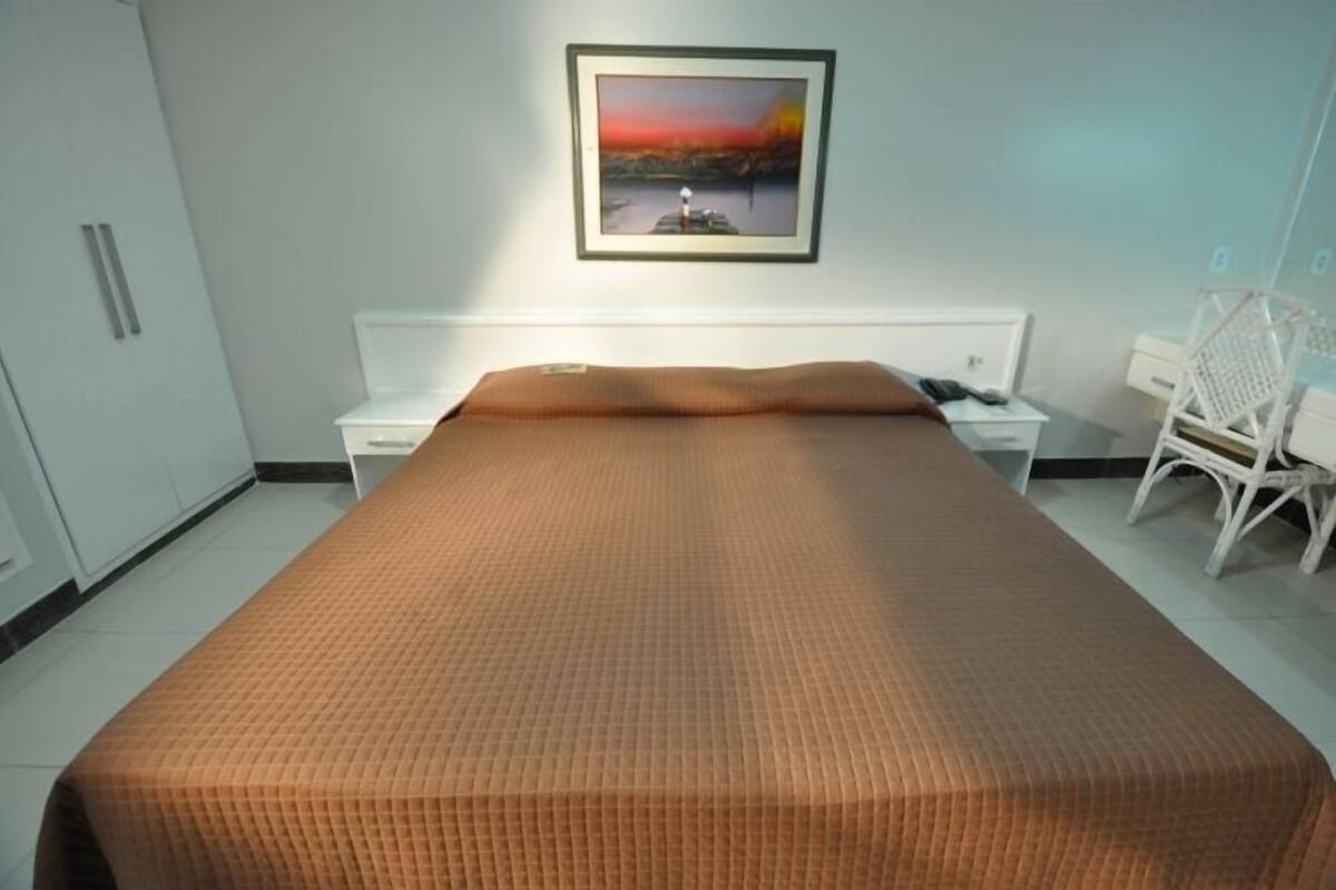 A hotel room with a large bed, a white nightstand, a chair, and a framed picture on the wall.
