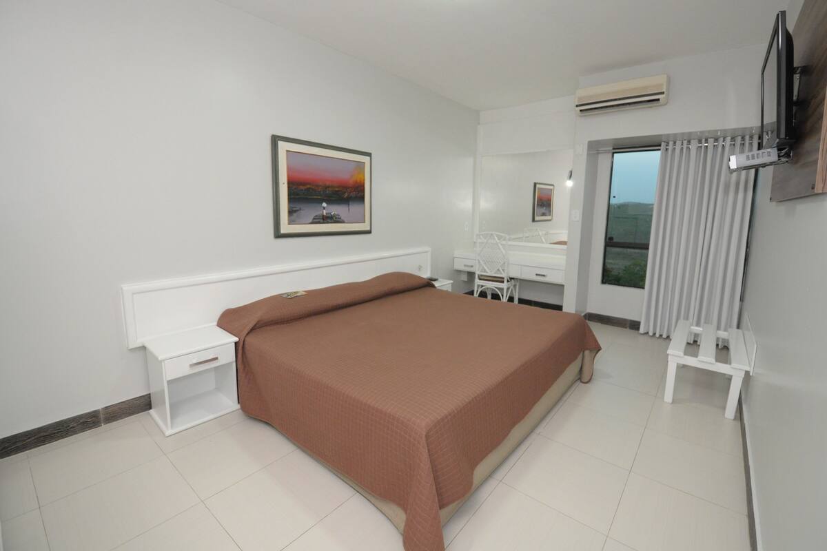 A bedroom with a bed, a desk, a chair, a TV, and a window with curtains.