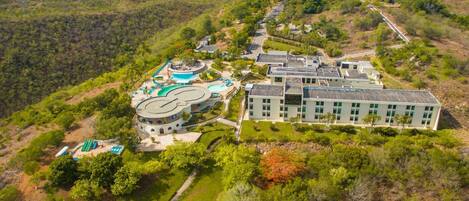 An aerial view of a resort with a large building, swimming pools, and surrounding greenery.