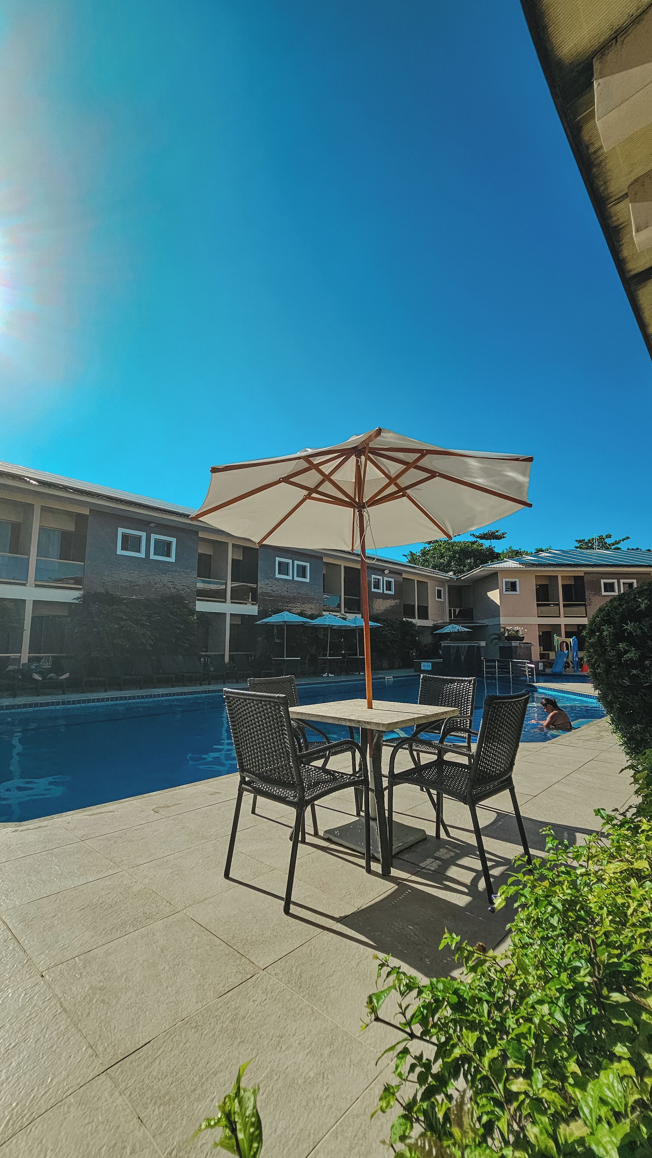 An outdoor pool area with a table and chairs, a large umbrella, and a building in the background.