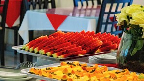 A buffet table with various fruit slices, a vase with yellow flowers, and a brick wall in the background. - Safira Praia Hotel (Porto Seguro)