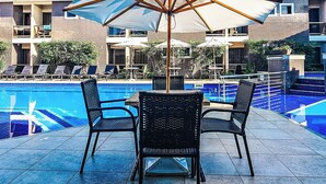 A poolside area with a table and chairs, umbrellas, and a view of a building with multiple balconies. - Safira Praia Hotel (Porto Seguro)