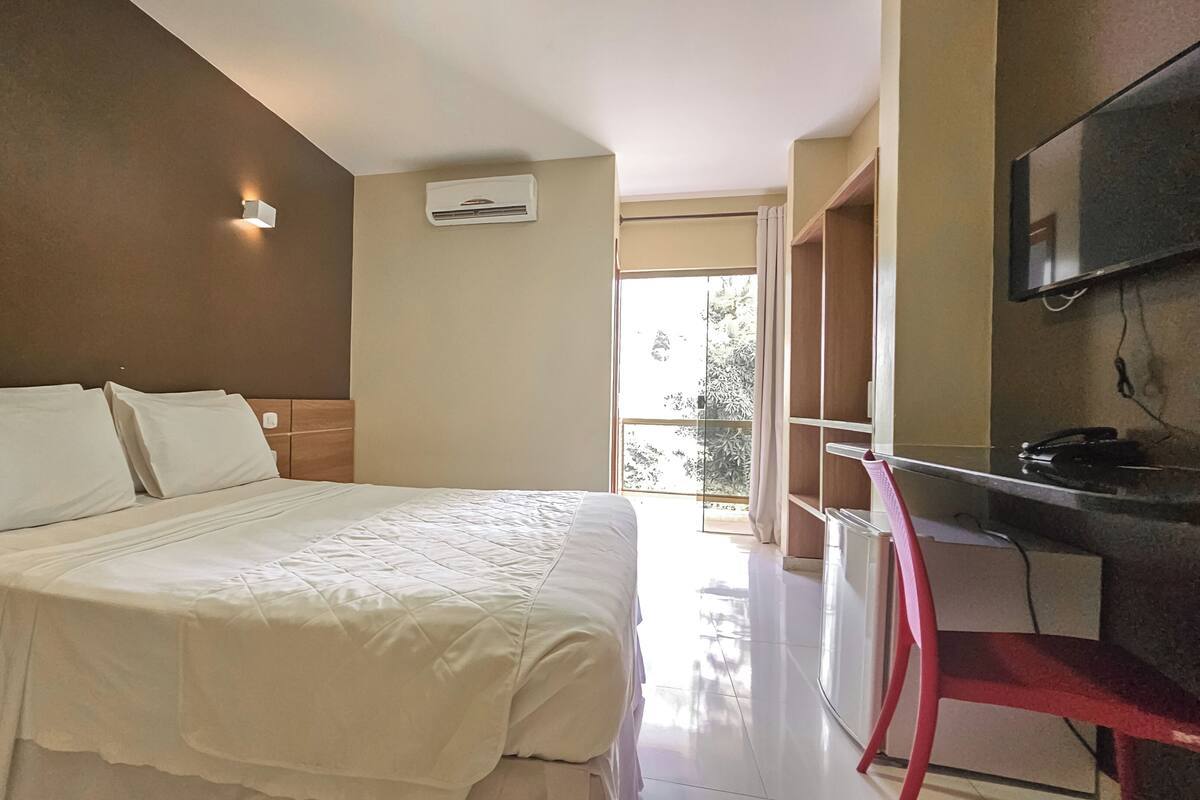 A hotel room with a bed, a desk, a chair, an air conditioning unit, and a TV mounted on the wall.