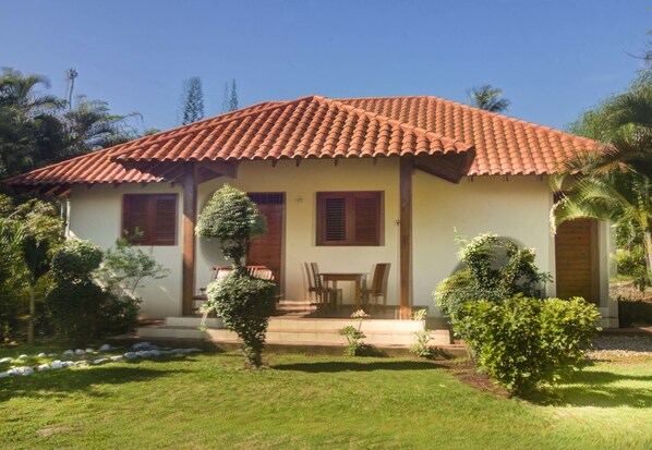 A two-story house with a red-tiled roof, a small porch with a table and chairs, and a well-maintained garden with various plants and a lawn.