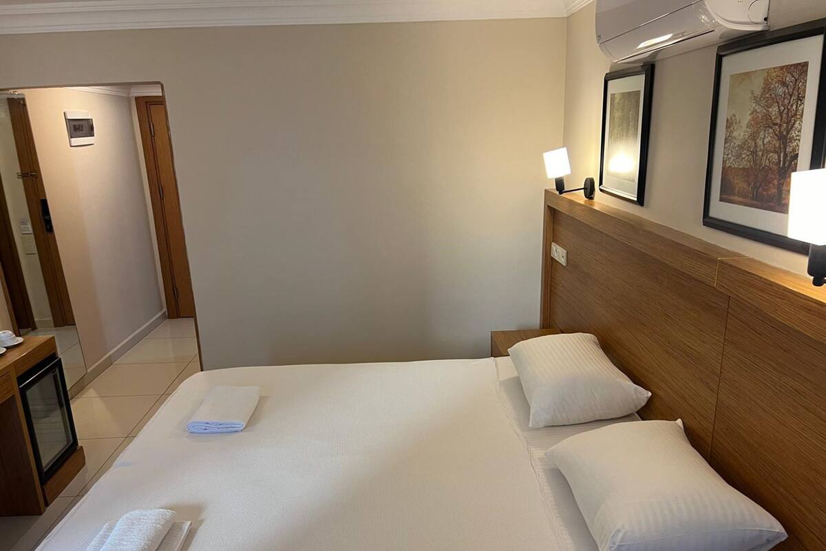 A hotel room with a large bed, two pillows, a bedside table with a lamp, a framed picture on the wall, and a door leading to another room.