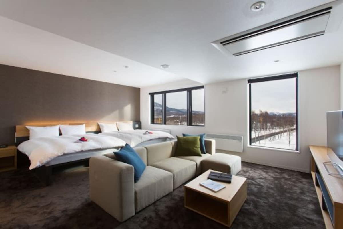 A modern hotel room with a bed, two sofas, a coffee table, a TV, and large windows offering a city view.