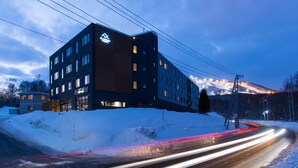 Un hotel moderno con varios pisos, un logotipo destacado en la fachada y un paisaje nevado al fondo.