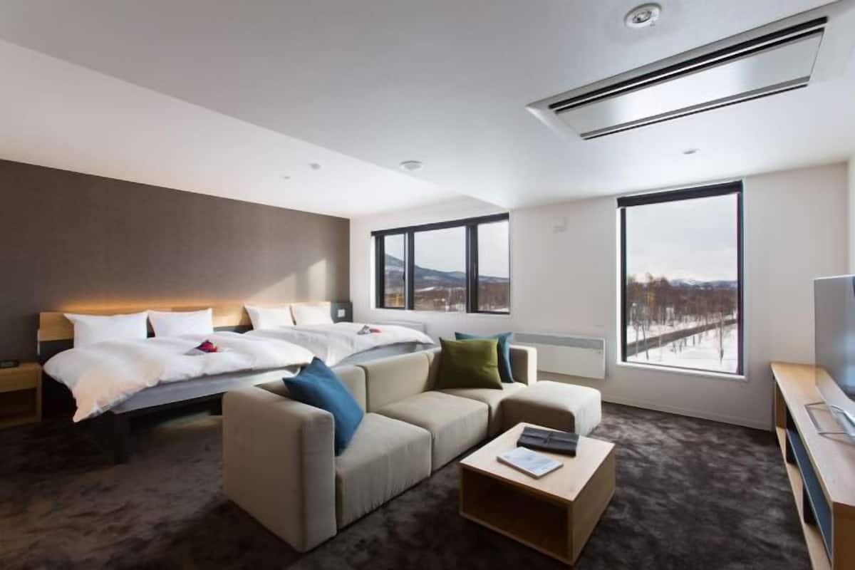 Habitación de hotel moderna con una cama, dos sofás, una mesita de centro, televisor y ventanas amplias con vistas a la ciudad.