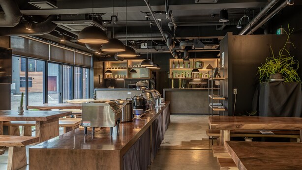 Un café moderno con mesas de madera, una barra alargada, ventanas amplias e iluminación de estilo industrial.