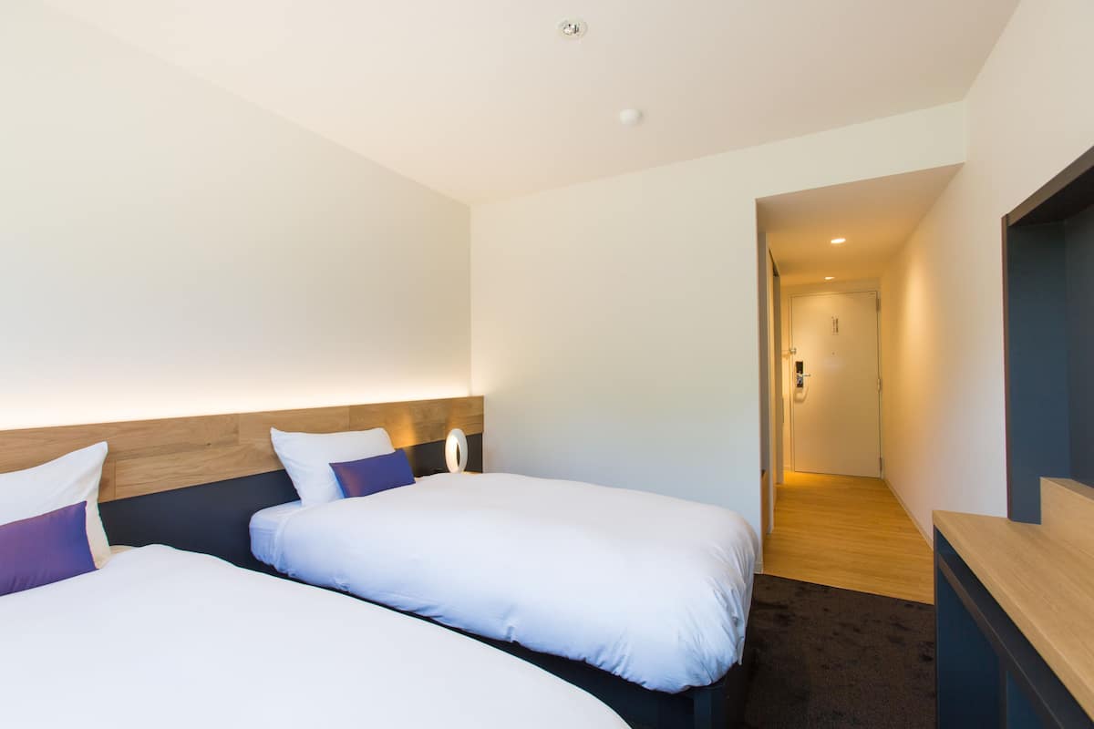 Habitación de hotel con dos camas, cabecero de madera, un escritorio pequeño y vista a un pasillo.