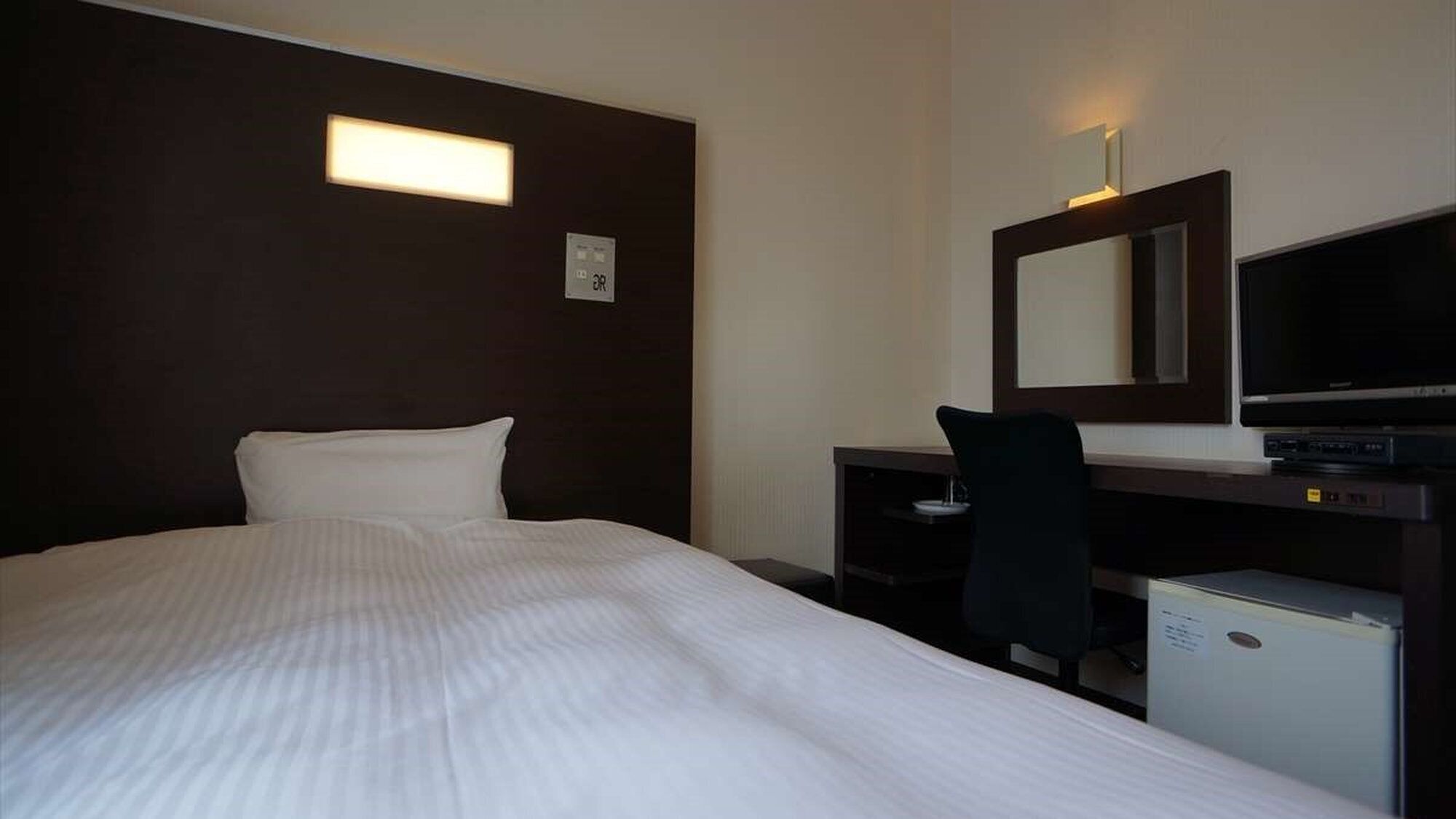A hotel room with a bed, a desk, a chair, a television, and a small refrigerator.