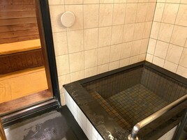 Área de chuveiro com azulejos, uma banheira preta, um banco de madeira e um painel de controle branco fixado na parede.