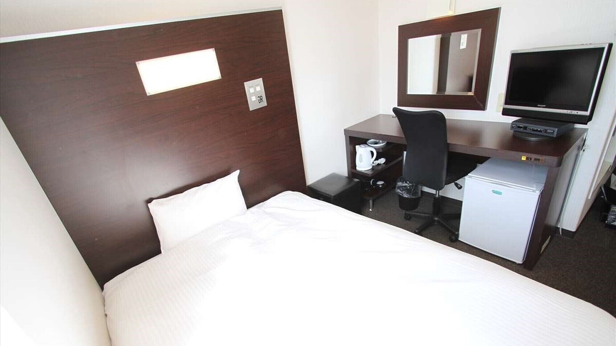 A hotel room with a bed, a desk with a computer, a chair, a small refrigerator, and a mirror.