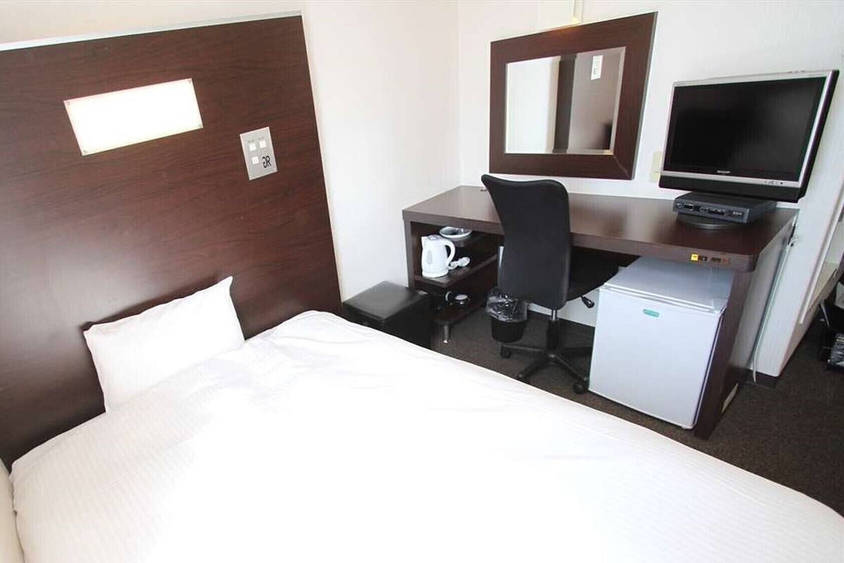 Een hotelkamer met een bed, een bureau met een computer, een stoel, een kleine koelkast en een spiegel.