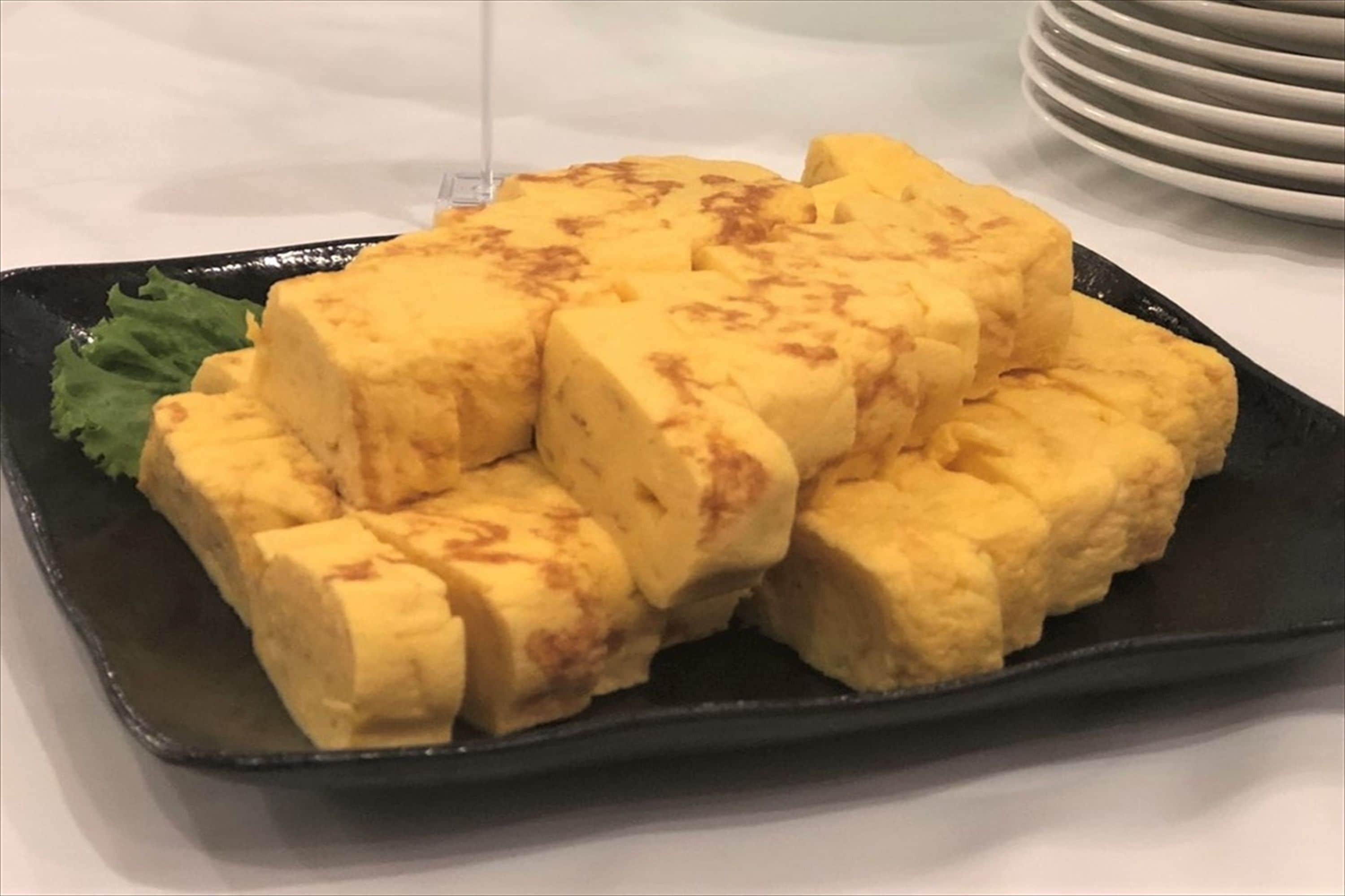 Une assiette rectangulaire noire sur laquelle sont disposés des cubes de tofu, formant une pyramide, et accompagnée d’une petite feuille verte.
