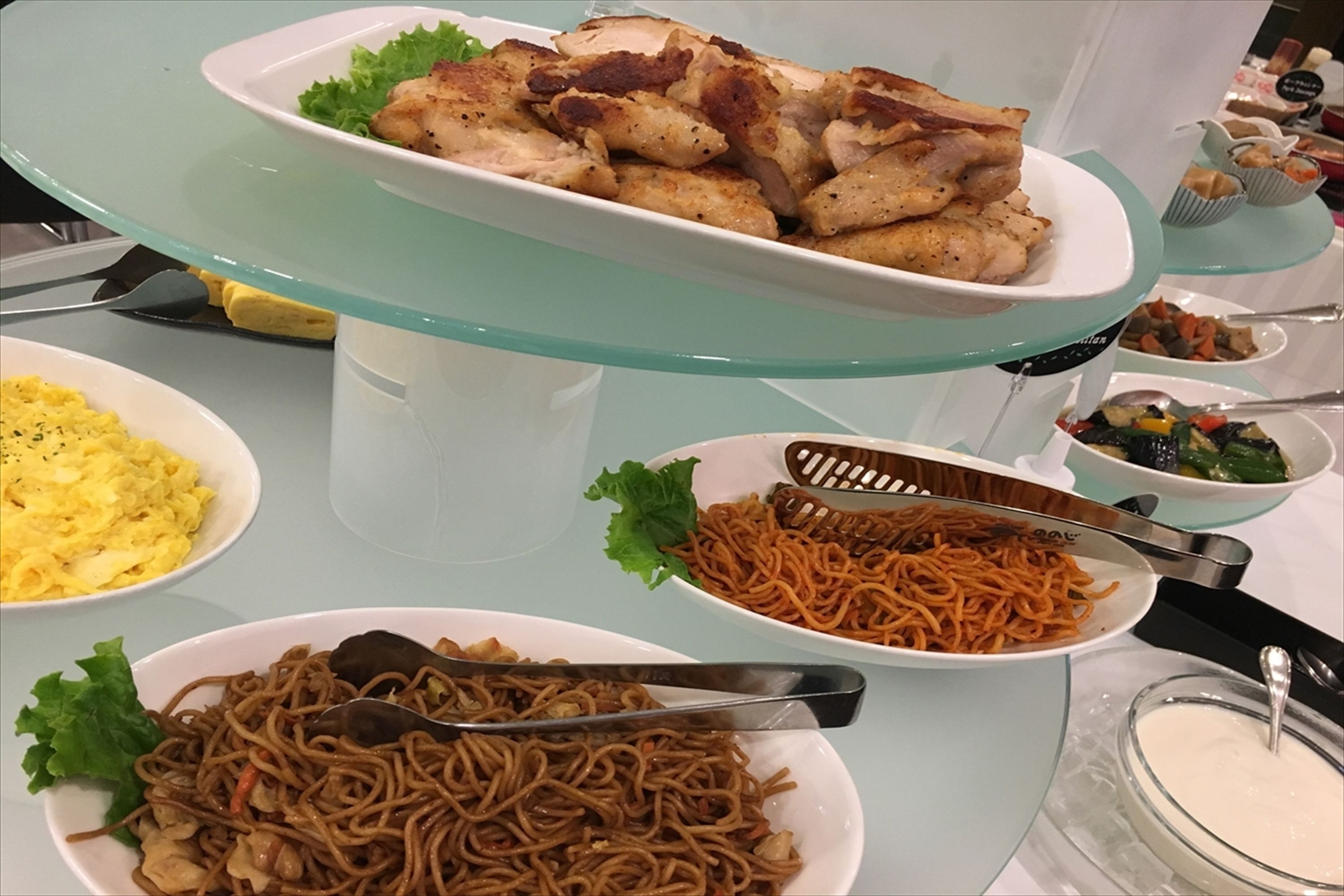 Una mesa de buffet con diversos platos, incluyendo pollo frito, fideos, huevos revueltos y verduras.