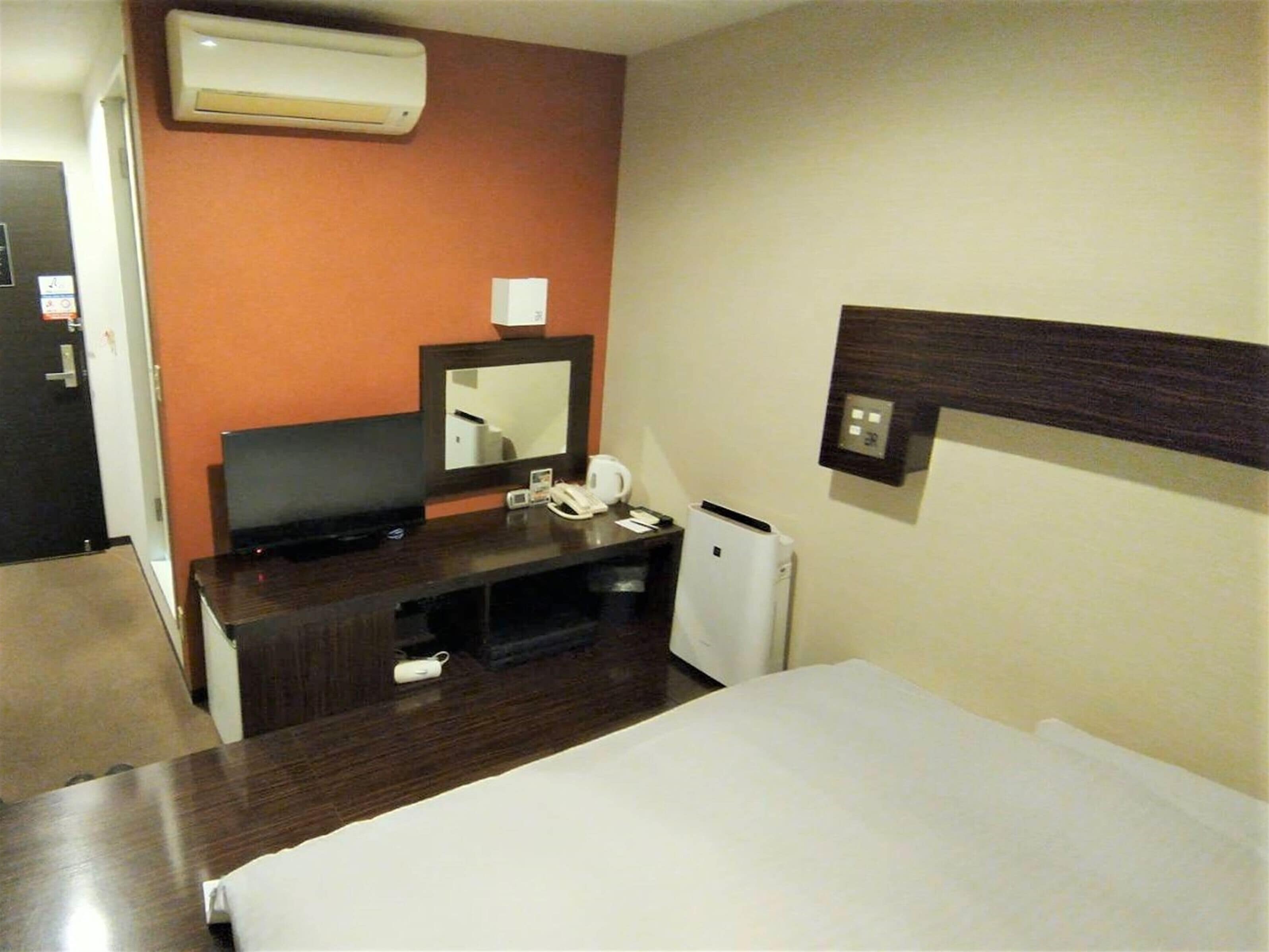 A hotel room with a bed, a desk with a telephone, a mirror, and an air conditioning unit.