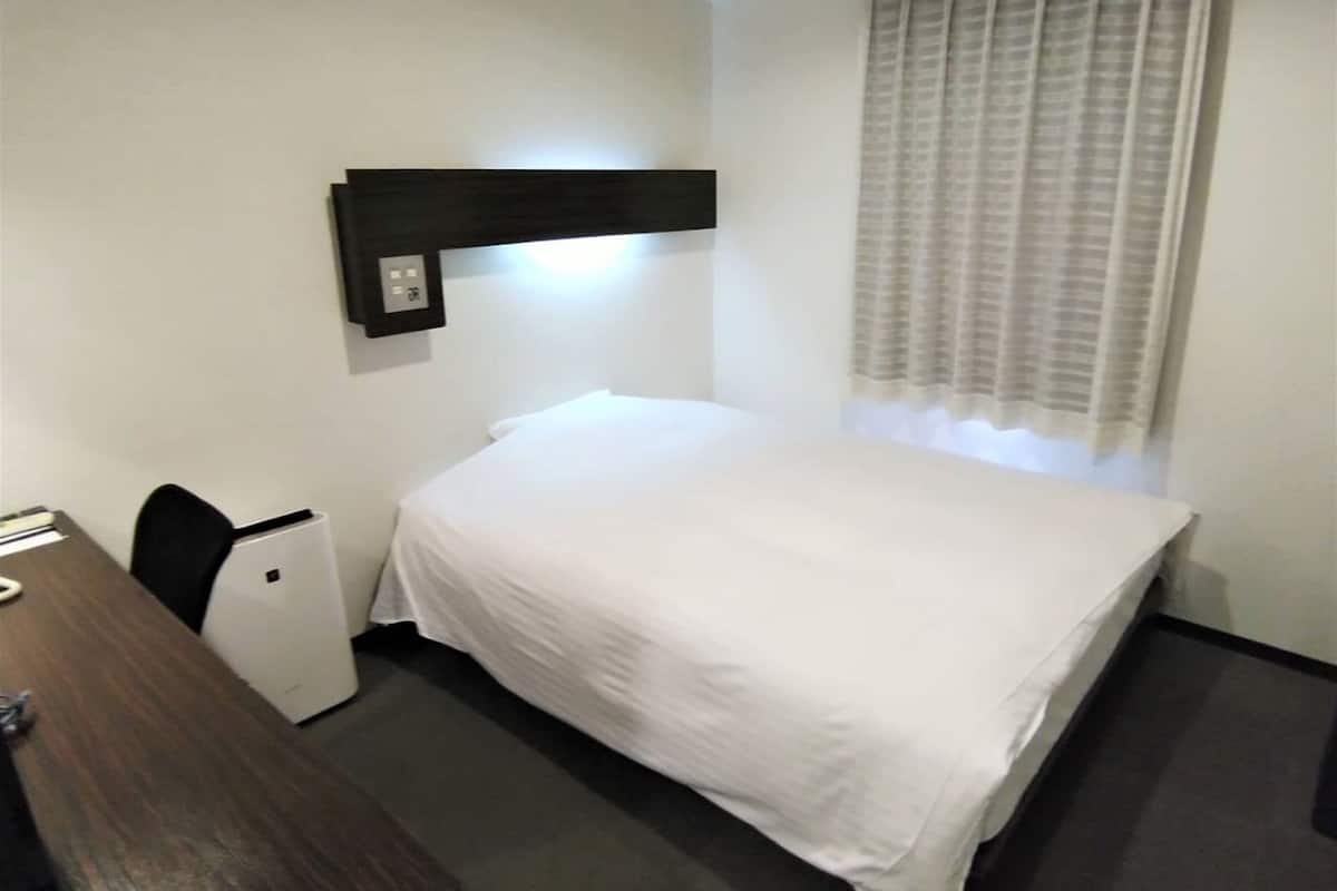 Une petite chambre d’hôtel, bien rangée, avec un lit simple, un bureau, une chaise et une fenêtre avec des rideaux.