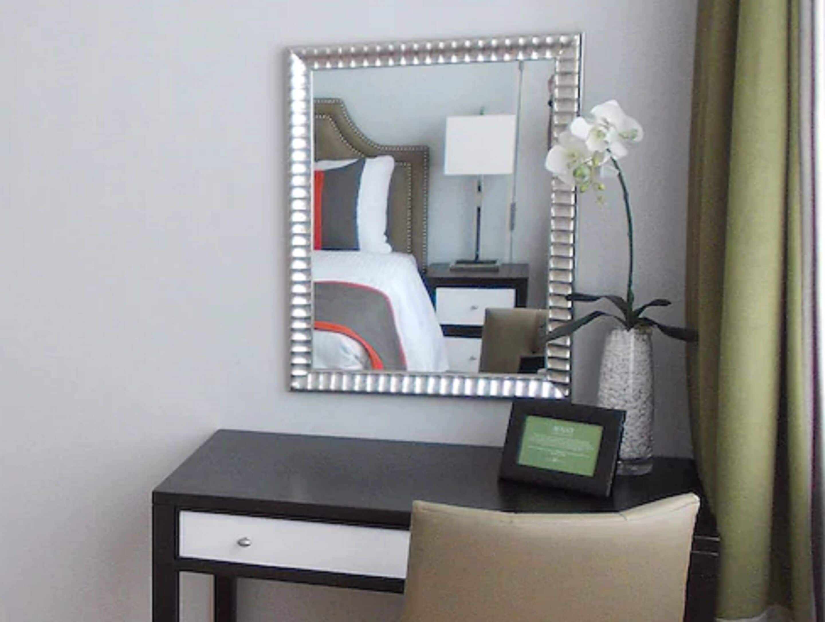 A hotel room with a bed, a bedside table with a lamp, a mirror, a vase with an orchid, and a desk with a chair.