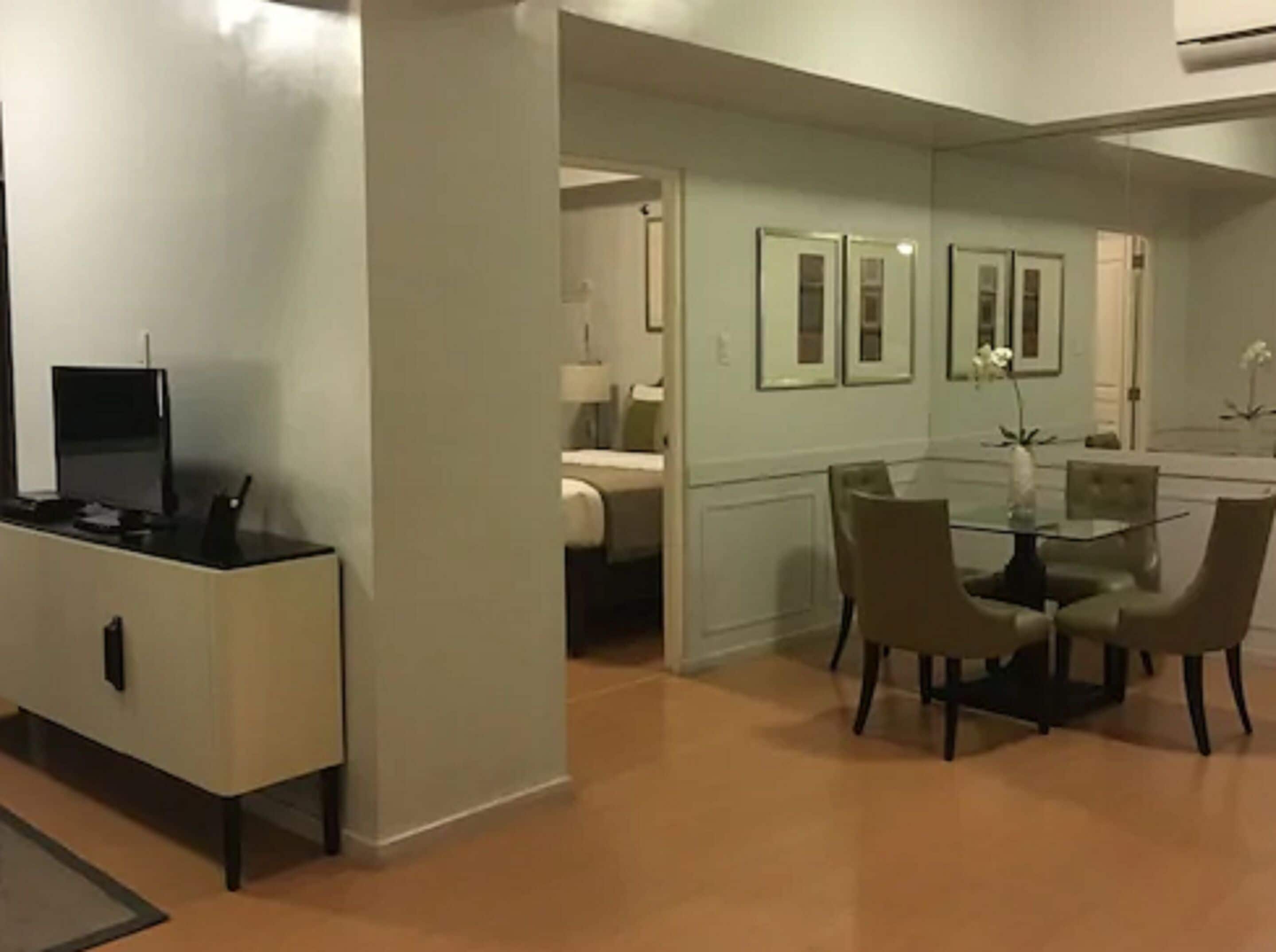 Een moderne hotelkamer met een eethoek, inclusief een glazen tafel en stoelen, een flatscreen televisie en een kitchenette.