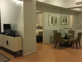 Ein modernes Hotelzimmer mit Essbereich, ausgestattet mit einem Glastisch und Stühlen, einem Flachbildfernseher und einer kleinen Küchenzeile.