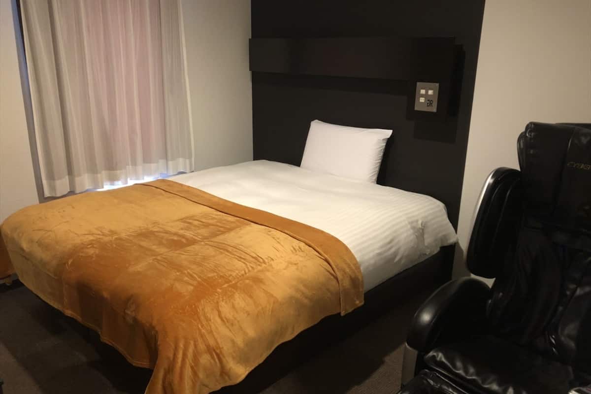 A hotel room with a bed, a black leather chair, and a wall-mounted control panel.
