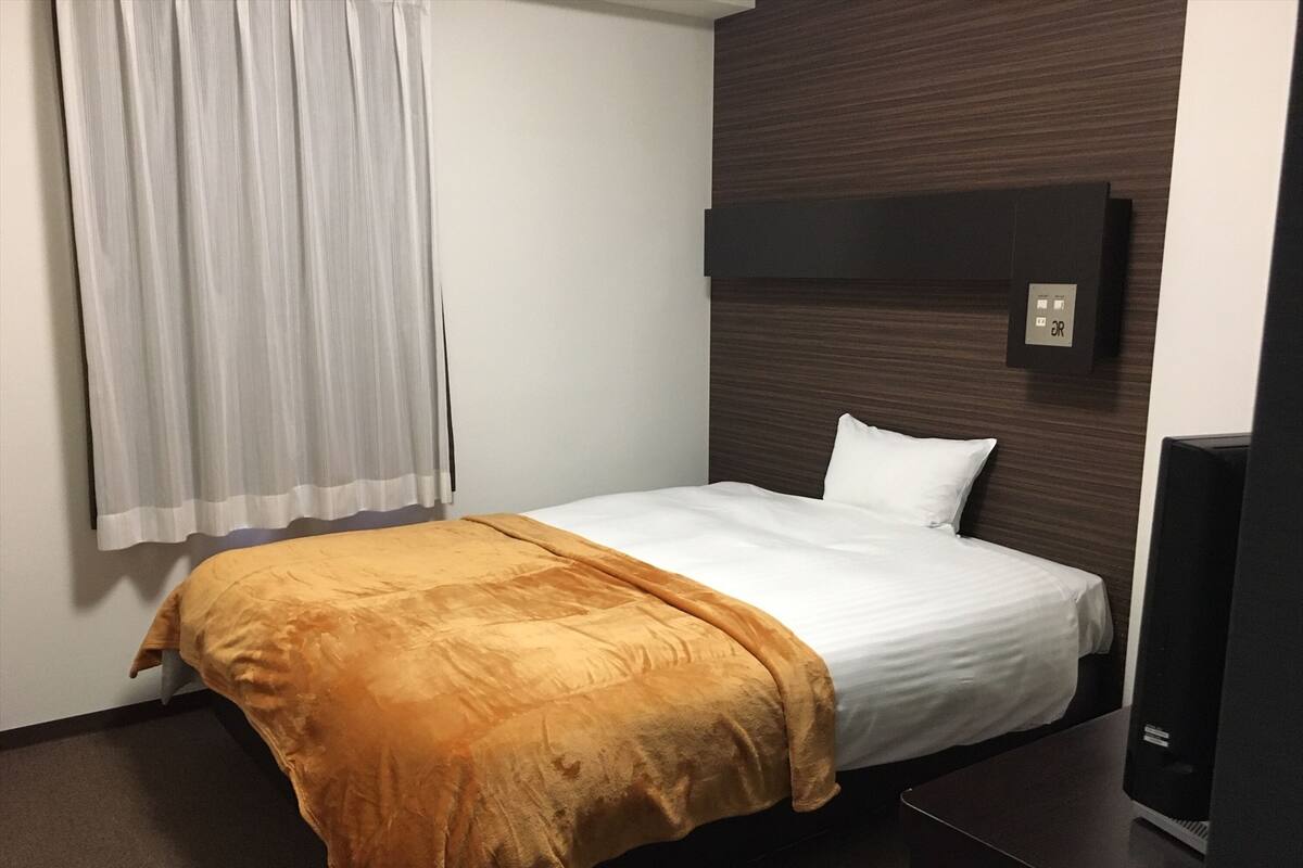 A hotel room with a single bed, a wooden headboard, a television, and a window with curtains.