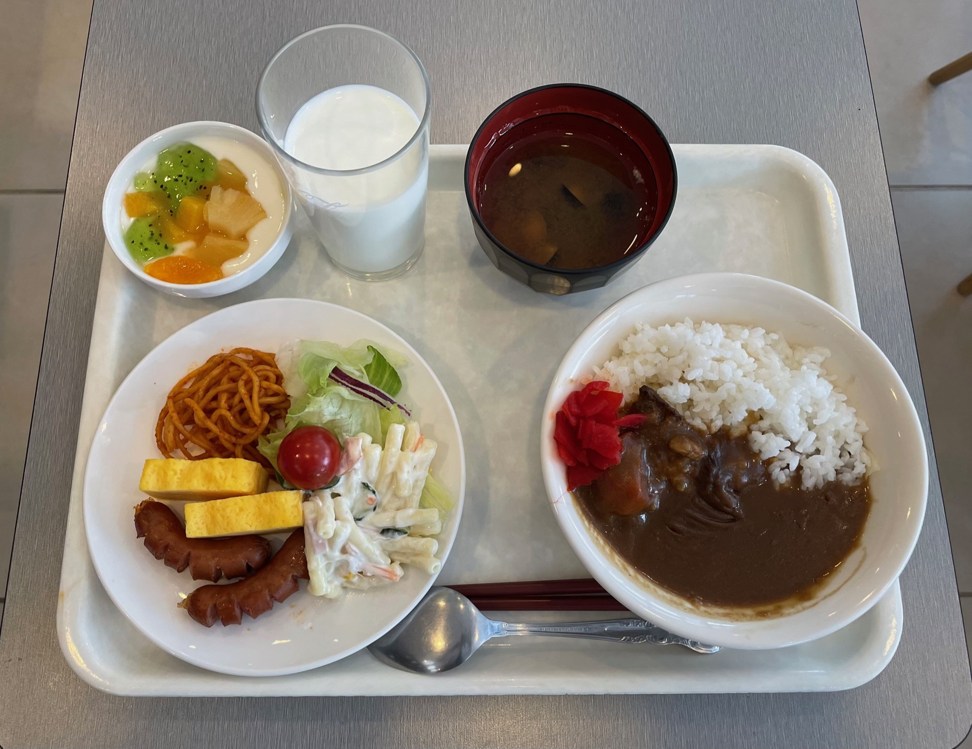 Una bandeja con un plato de fideos, ensalada, tofu y salchicha; un tazón de arroz con salsa; un vaso de leche; y un tazón de sopa.
