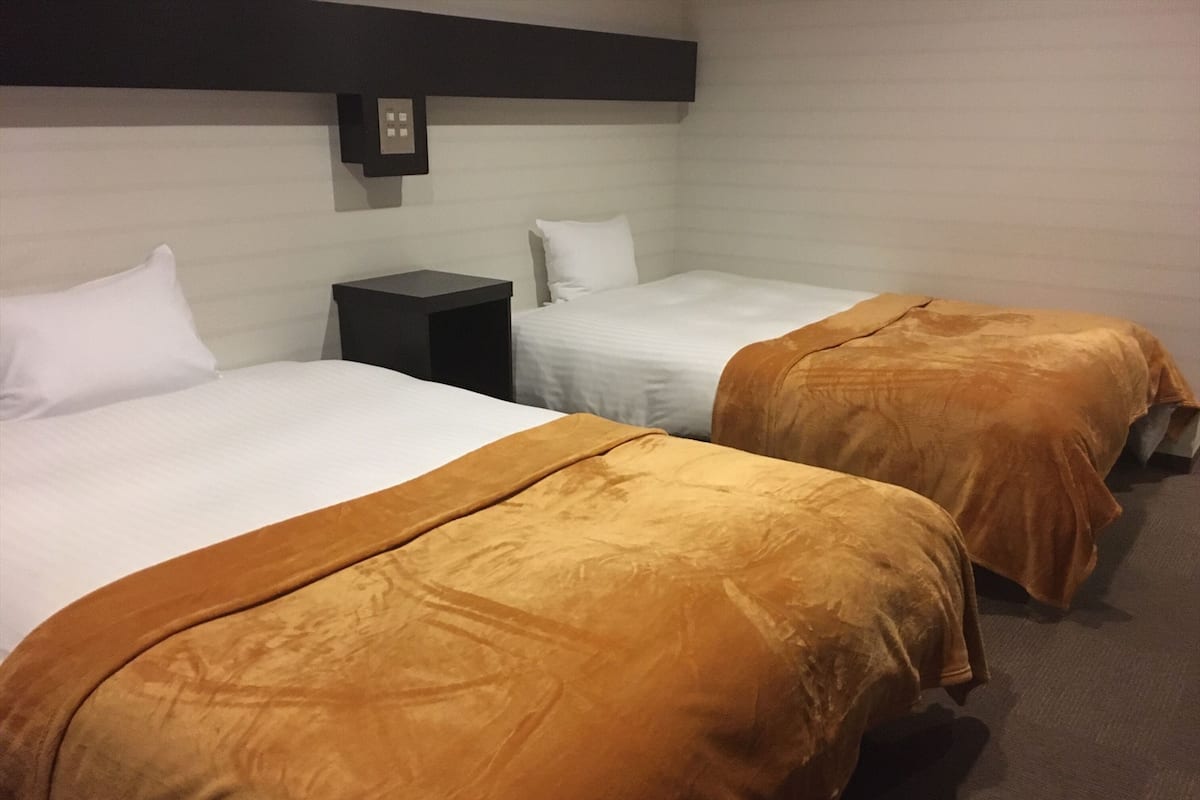 Two single beds with white and brown bedding, a small bedside table, and a wall-mounted light fixture.