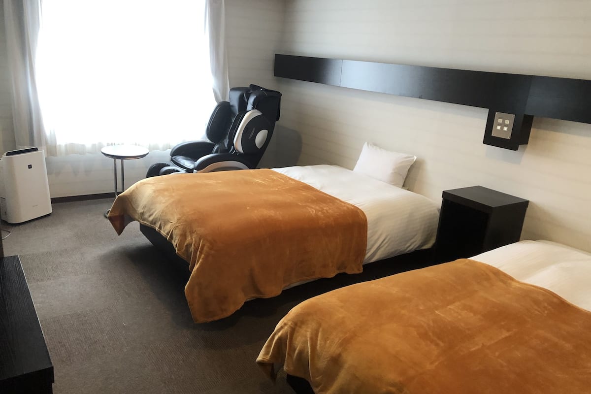 A hotel room with two beds, a massage chair, a small table, and a wall-mounted TV.