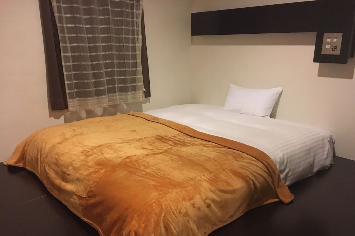A neatly made bed with a white and orange bedspread, a plain wall, and a window with a checkered curtain.