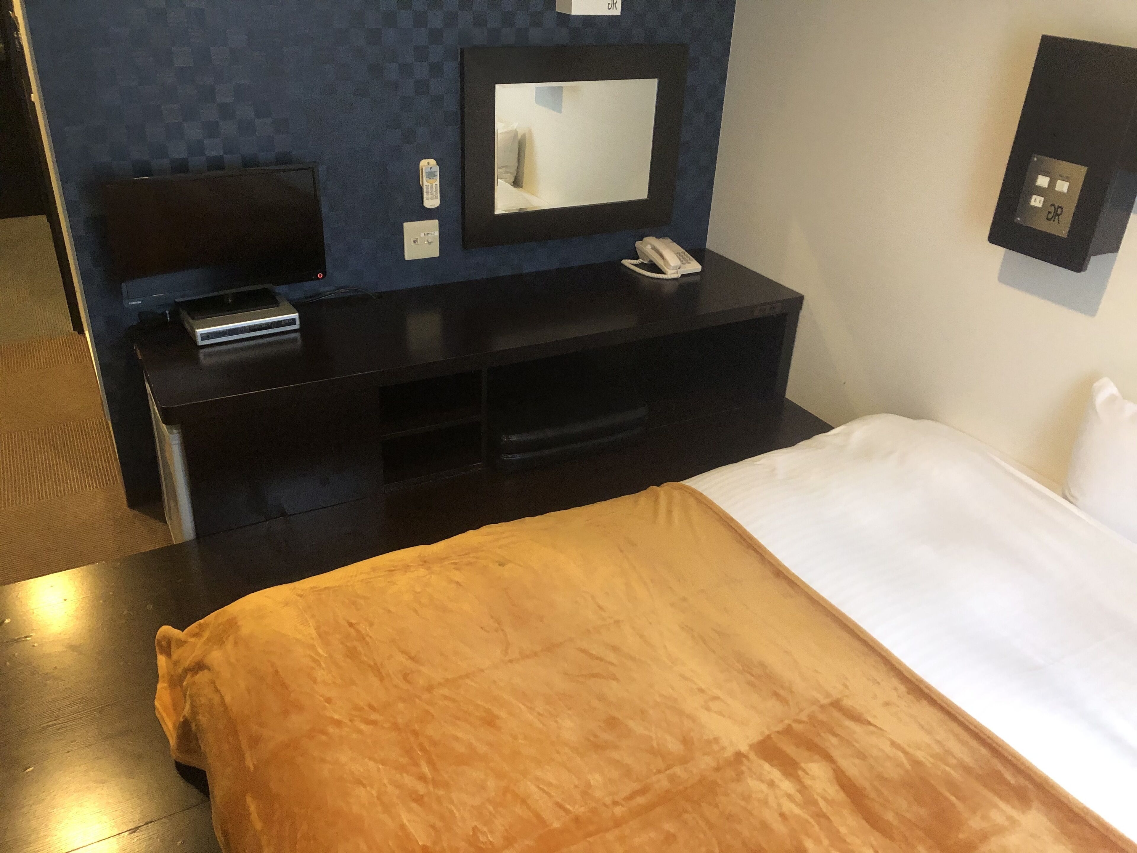 Et hotelværelse med seng, fjernsyn på fod, telefon og kontrolpanel på væggen.