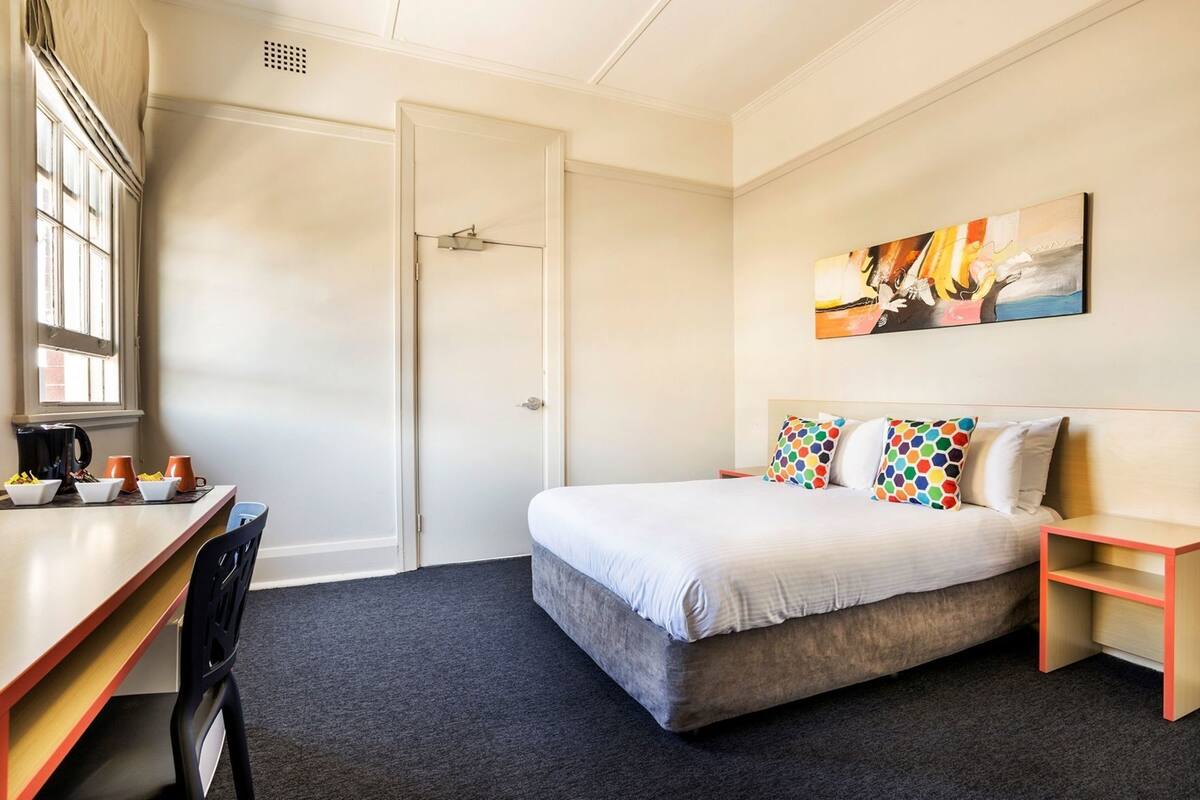Habitación de hotel con cama, escritorio con silla, cafetera y un cuadro en la pared.