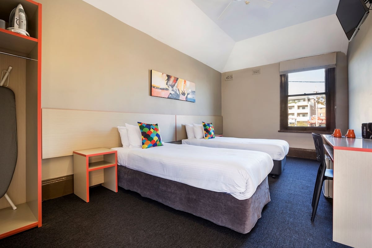 Habitación de hotel con dos camas, un escritorio, una silla, una repisa con una tetera y un cuadro en la pared.
