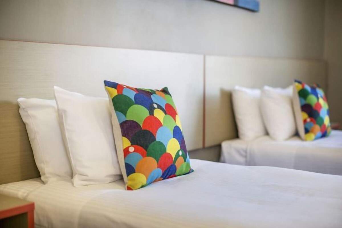 Ein Hotelzimmer mit zwei Betten, jeweils mit weißer Bettwäsche und bunten, gemusterten Kissen.