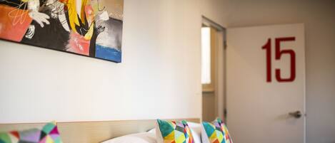 Un cuarto de hotel con una cama, almohadas coloridas, un cuadro en la pared y una puerta con el número 15.