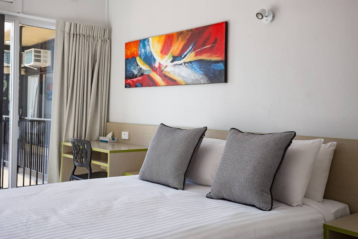 A hotel room with a bed, two pillows, a painting on the wall, a window with curtains, and a small table with a chair.