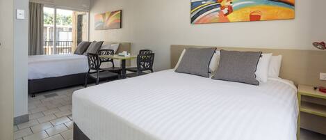 Quarto de hotel com duas camas, uma escrivaninha e uma cadeira. Há um quadro na parede e uma janela com cortinas.