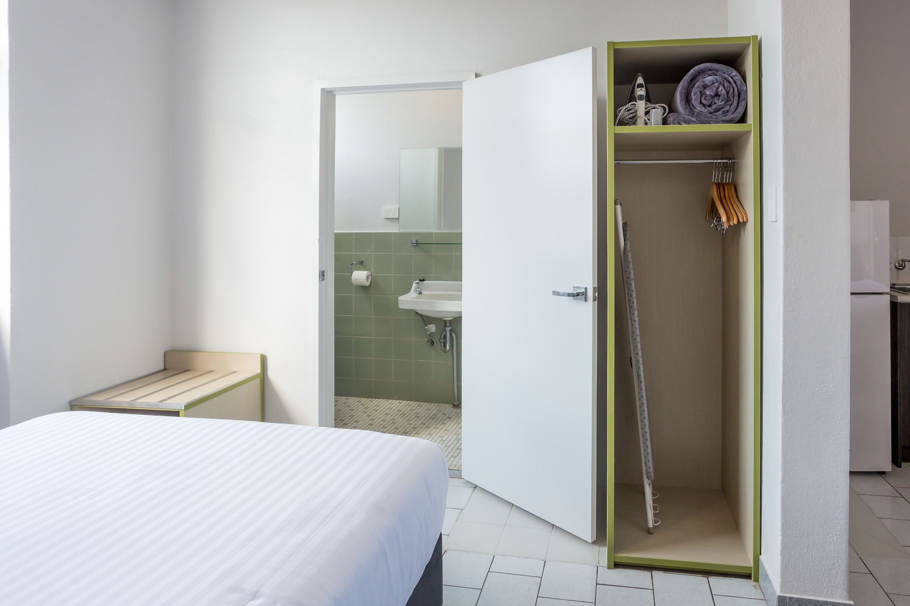 A hotel room with a bed, a small bedside table, a wardrobe with a mirror, and a bathroom visible through an open door.