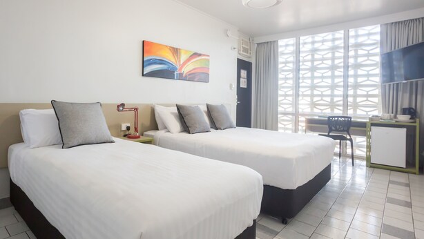 Habitación de hotel con dos camas, un escritorio, una silla y una ventana con un diseño geométrico.