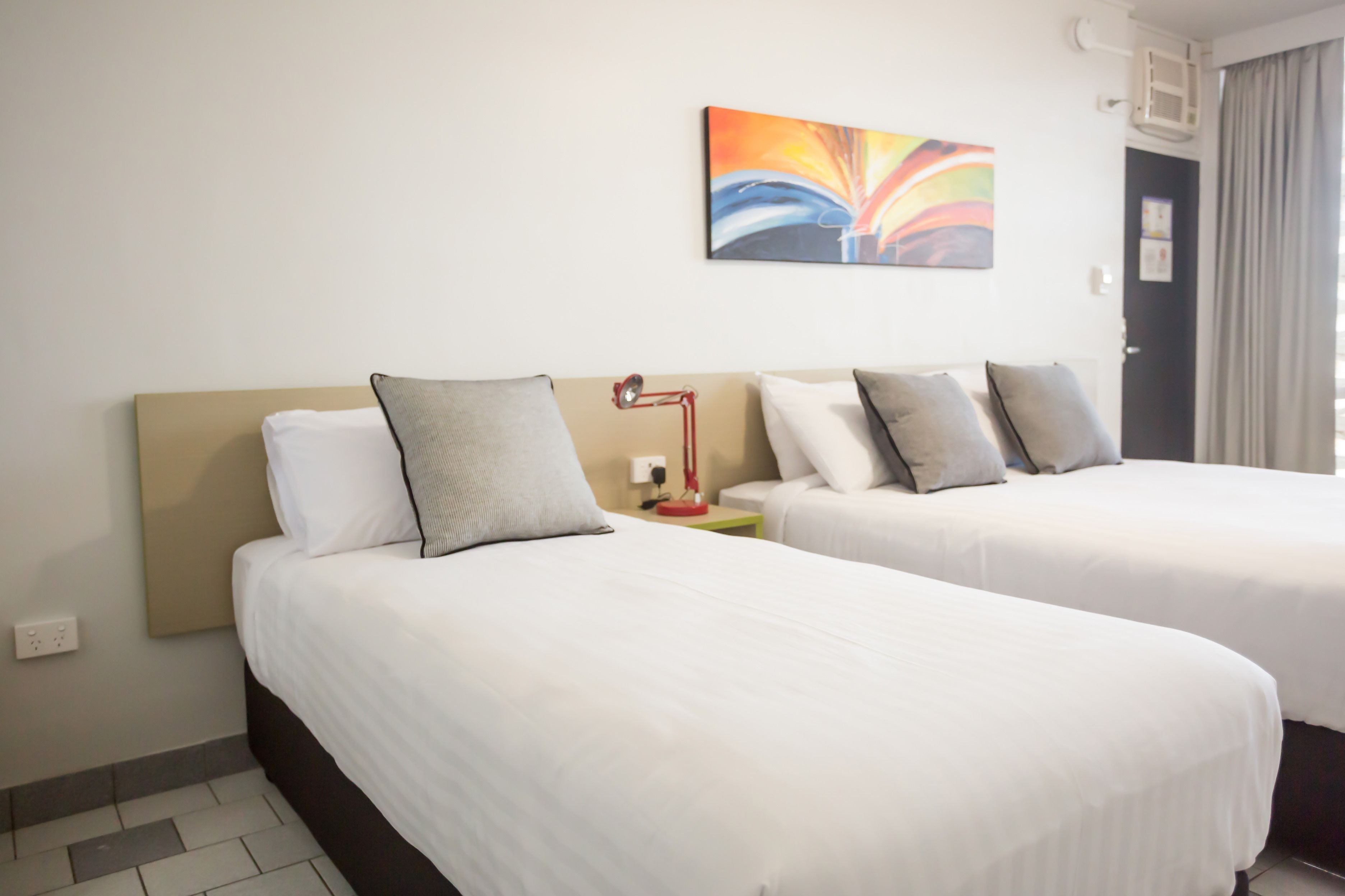 Ein Hotelzimmer mit zwei Betten, einem modernen abstrakten Gemälde, einer roten Lampe und einem Fenster mit Vorhängen.