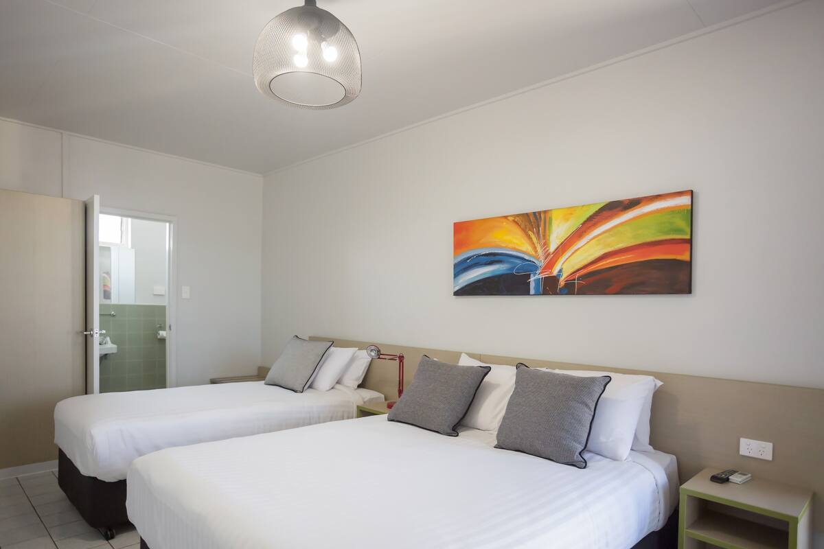 A hotel room with two beds, a painting on the wall, a bedside table with a lamp, and a door leading to a bathroom.