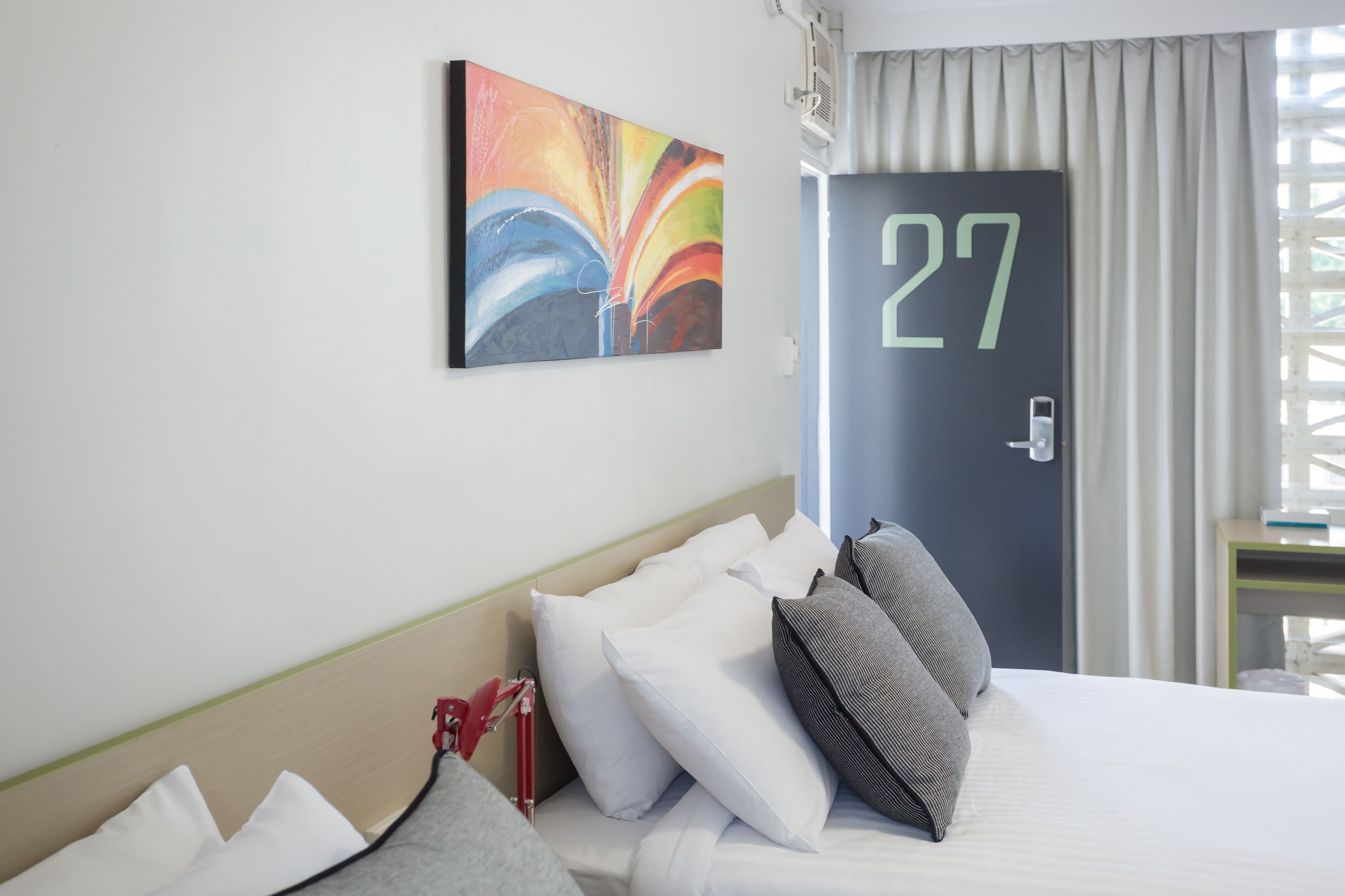 Una habitació d'hotel amb un llit, almoines, un quadre abstracte i colorit, una porta amb el número 27 i una finestra amb cortines.