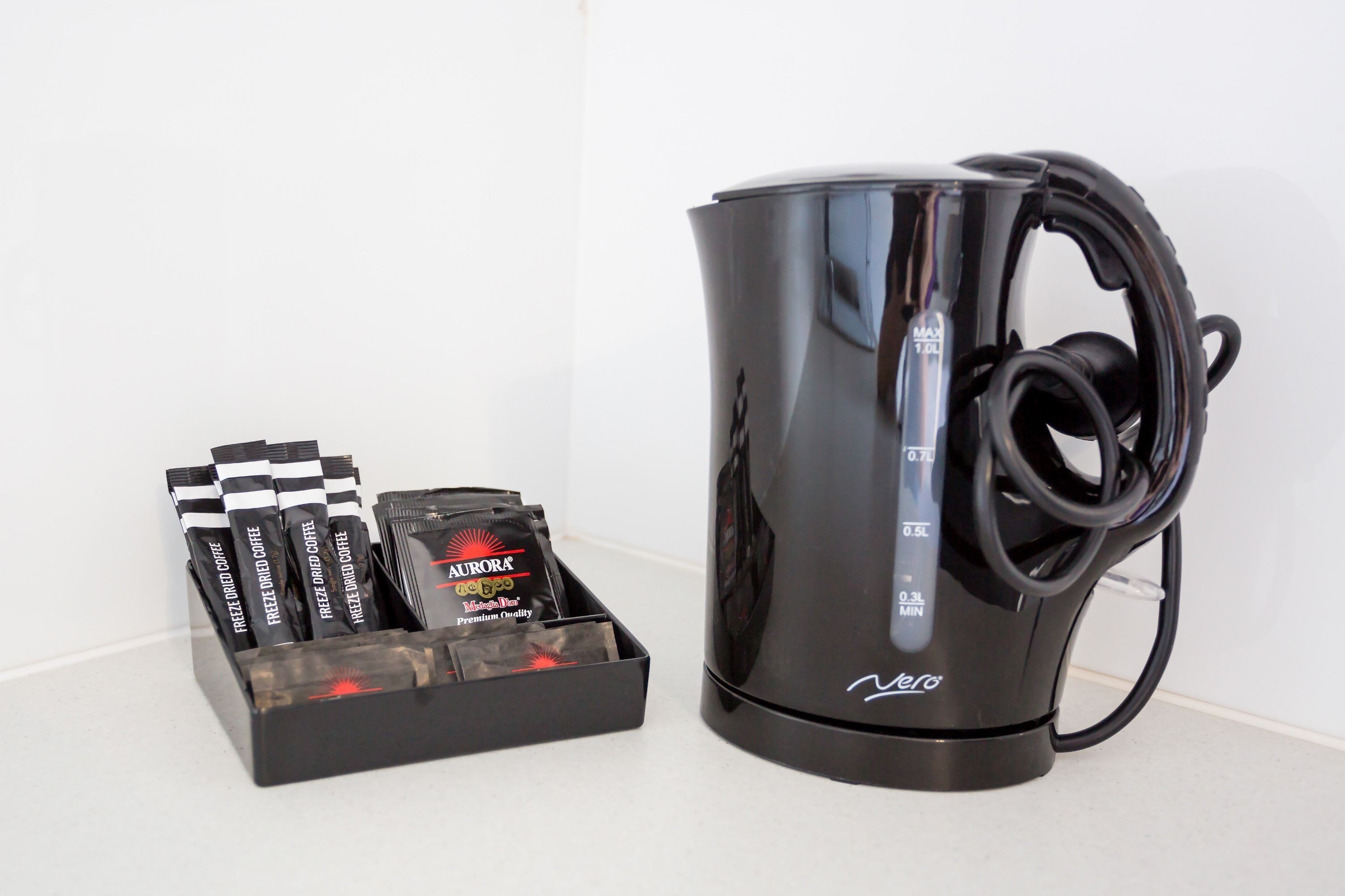 Una cafetera negra con asa y un cable de carga, junto a una caja con cápsulas de café y una taza de café con el logo de la marca.