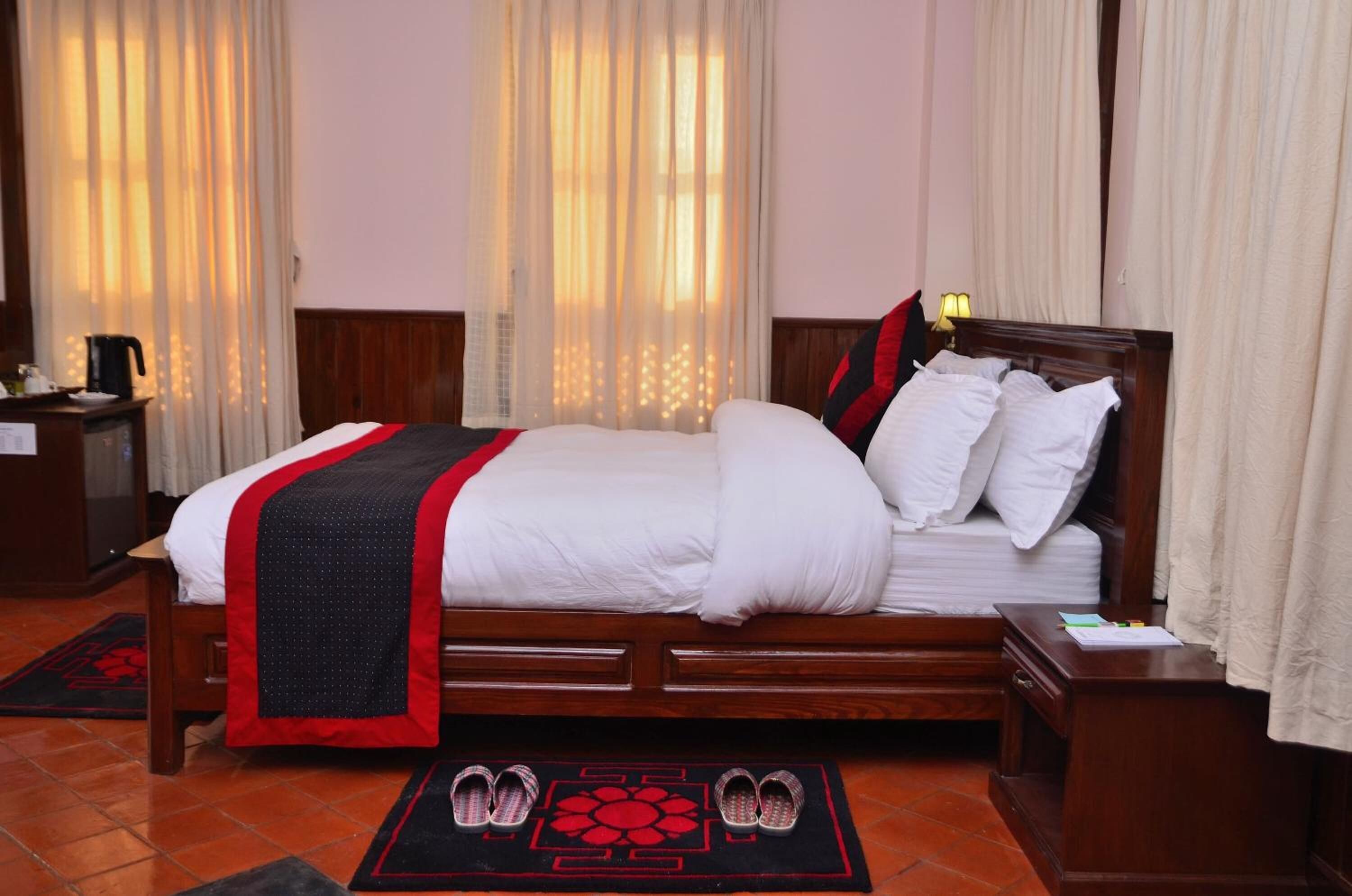 Una cama bien hecha con ropa blanca y una colcha rojo y negro, una mesita de noche de madera con una lámpara, y un par de zapatillas en un felpudo rojo y negro.