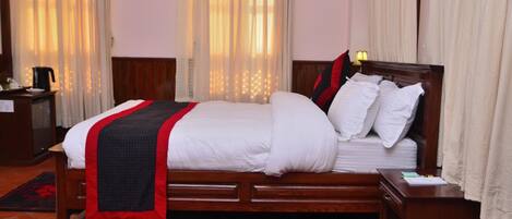 Una cama bien hecha con ropa blanca y una colcha rojo y negro, una mesita de noche de madera con una lámpara, y un par de zapatillas en un felpudo rojo y negro.