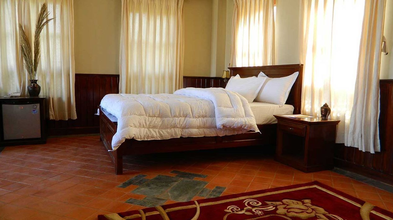 A neatly made double bed with a wooden headboard, a bedside table, and a nightstand with a lamp. A decorative vase with dried plants is on the nightstand.