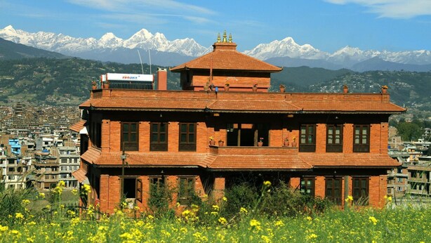 Ein traditionelles Backsteingebäude mit flachem Dach und kleinem Turm, umgeben von einem Feld gelber Blumen vor einer Bergkulisse.